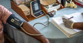 A health worker measures an adult’s blood pressure using a digital monitor, with the reading being recorded in a notebook.