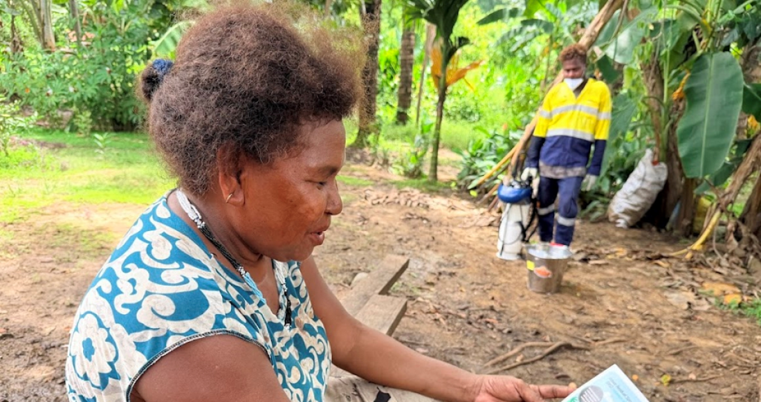 A person seated outdoors in Guadalcanal, Solomon Islands holds and reads an illustrated health information leaflet while a nearby community health worker stands on a dirt path surrounded by greenery, conducting community-based interventions.