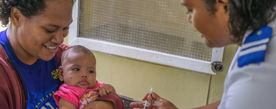 A health worker holds a syringe while preparing to administer a vaccine to a baby seated on a caregiver’s lap in a clinic setting.