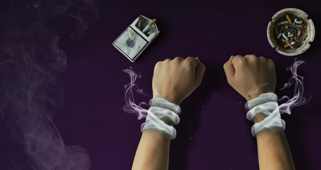 Two human wrists wrapped tightly with white bands formed from the smoke of an ashtray with cigarette butts and an open cigarette pack are placed on a surface.